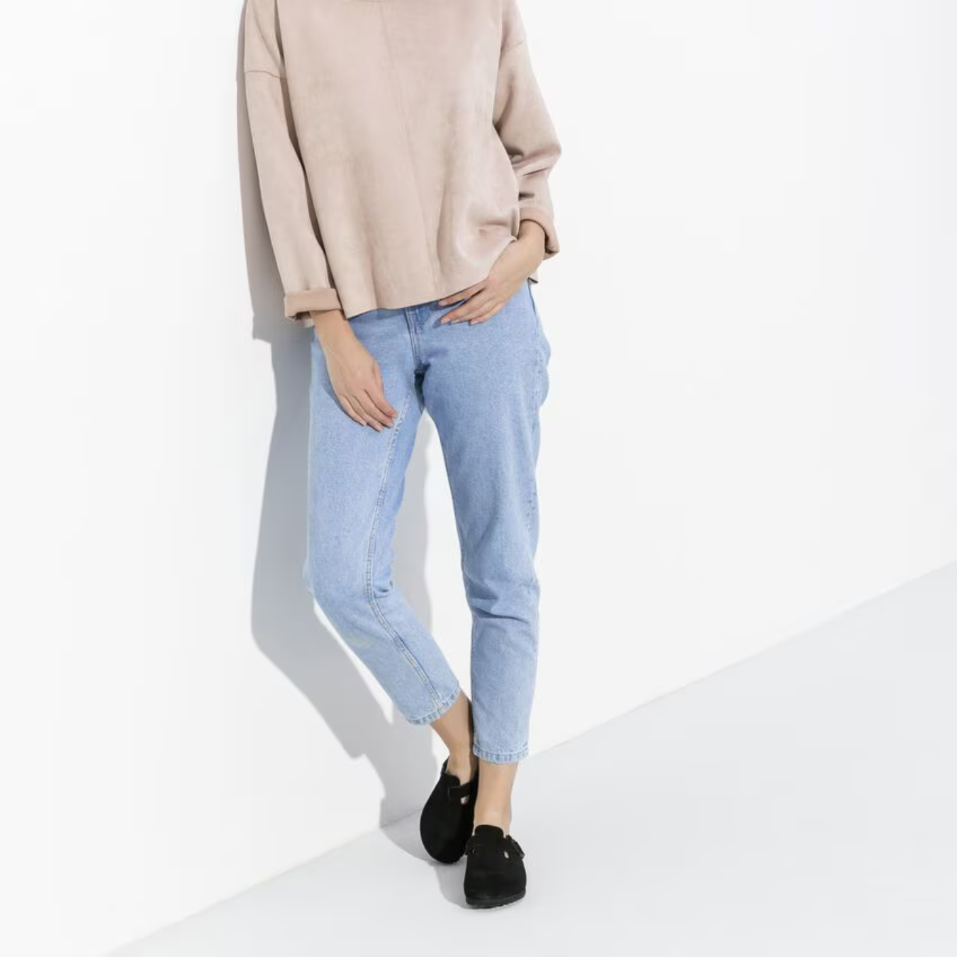 Kvinde iført beige sweater og lyseblå jeans, læner sig mod hvid væg. Mode, afslappet stil, komfortabelt tøj, minimalistisk look, sort fodtøj.