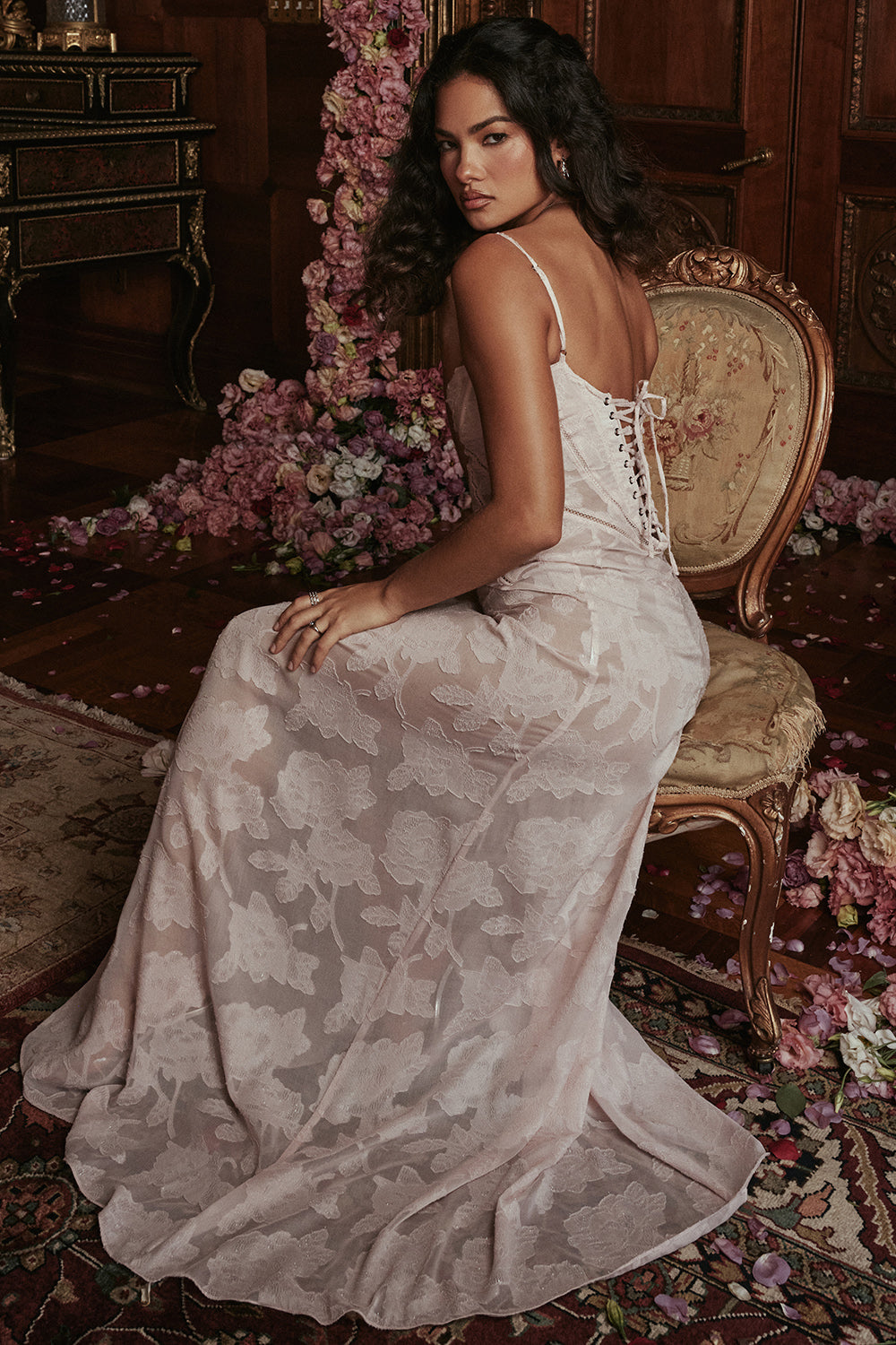 Kvinde i elegant, hvid blondekjole sidder på antik stol i overdådigt dekoreret rum med blomster. Bryllupsmode, luksus, romantisk atmosfære.