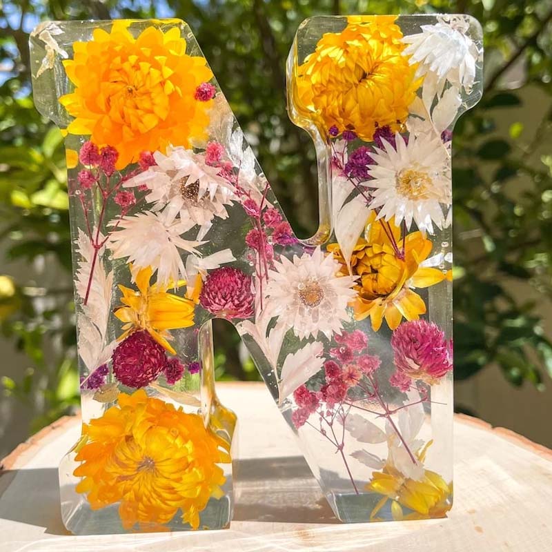 Bogstavet "N" i harpiks med indlejrede tørrede blomster, herunder gule og pink blomster, mod en grøn baggrund. Dekorativt og unikt design.