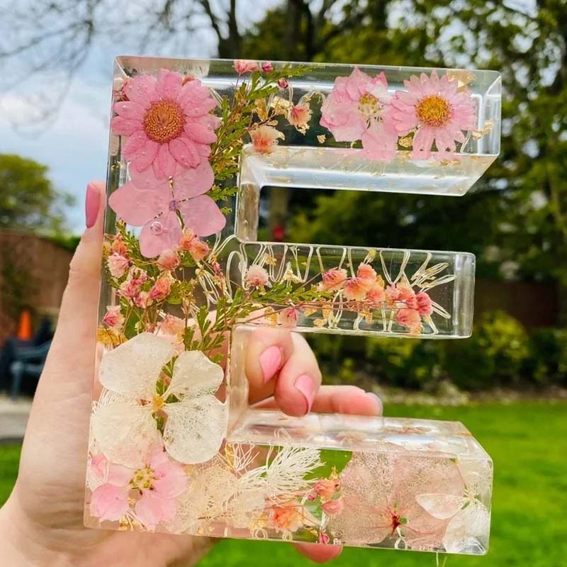 Hånd holder gennemsigtig harpiksbogstav "E" med indlejrede lyserøde blomster, udendørs baggrund. Unikt håndlavet dekorativt element.