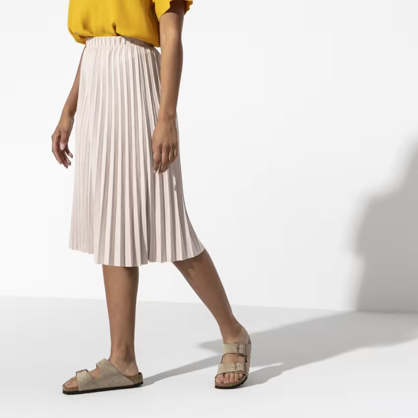 Person iført gul bluse, beige plisseret nederdel og beige sandaler. Mode, sommeroutfit, stilfuldt tøj, kvinde, casual look.
