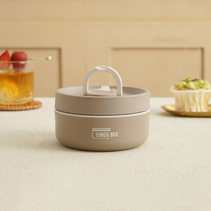 Beige madkasse med håndtag på hvidt bord, omgivet af en drink med frugt og en muffin. Perfekt til opbevaring af frokost.