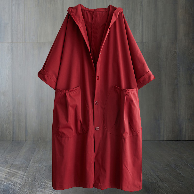 Rød regnfrakke med hætte og store lommer, hængende mod en grå trævæg. Stilfuldt og funktionelt design til regnfulde dage.