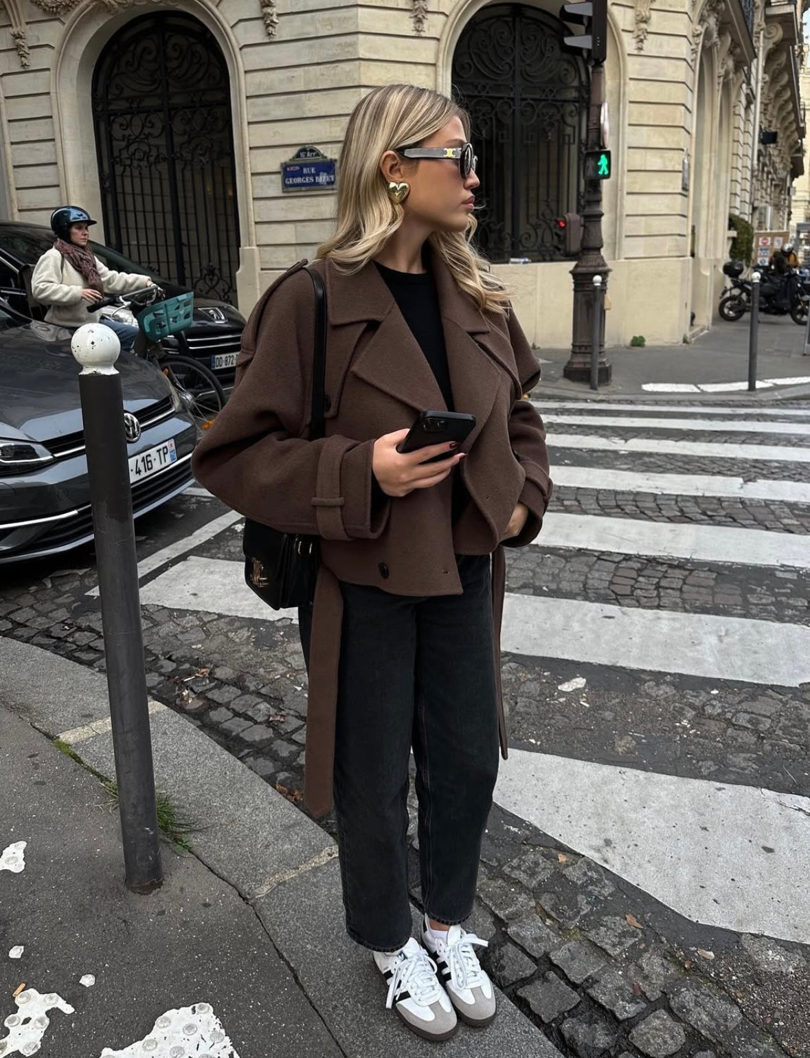 Kvinde i brun frakke og solbriller står ved fodgængerfelt i bymiljø, holder smartphone. Mode, gade, stil, efterårstøj, urbant look.