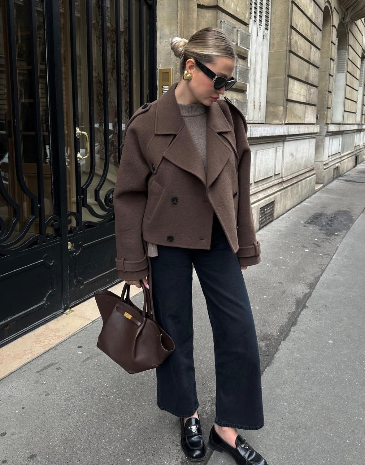 Kvinde i brun frakke og sorte bukser står på gaden med en brun taske. Hun bærer solbriller og loafers. Mode, gade, efterårstøj.