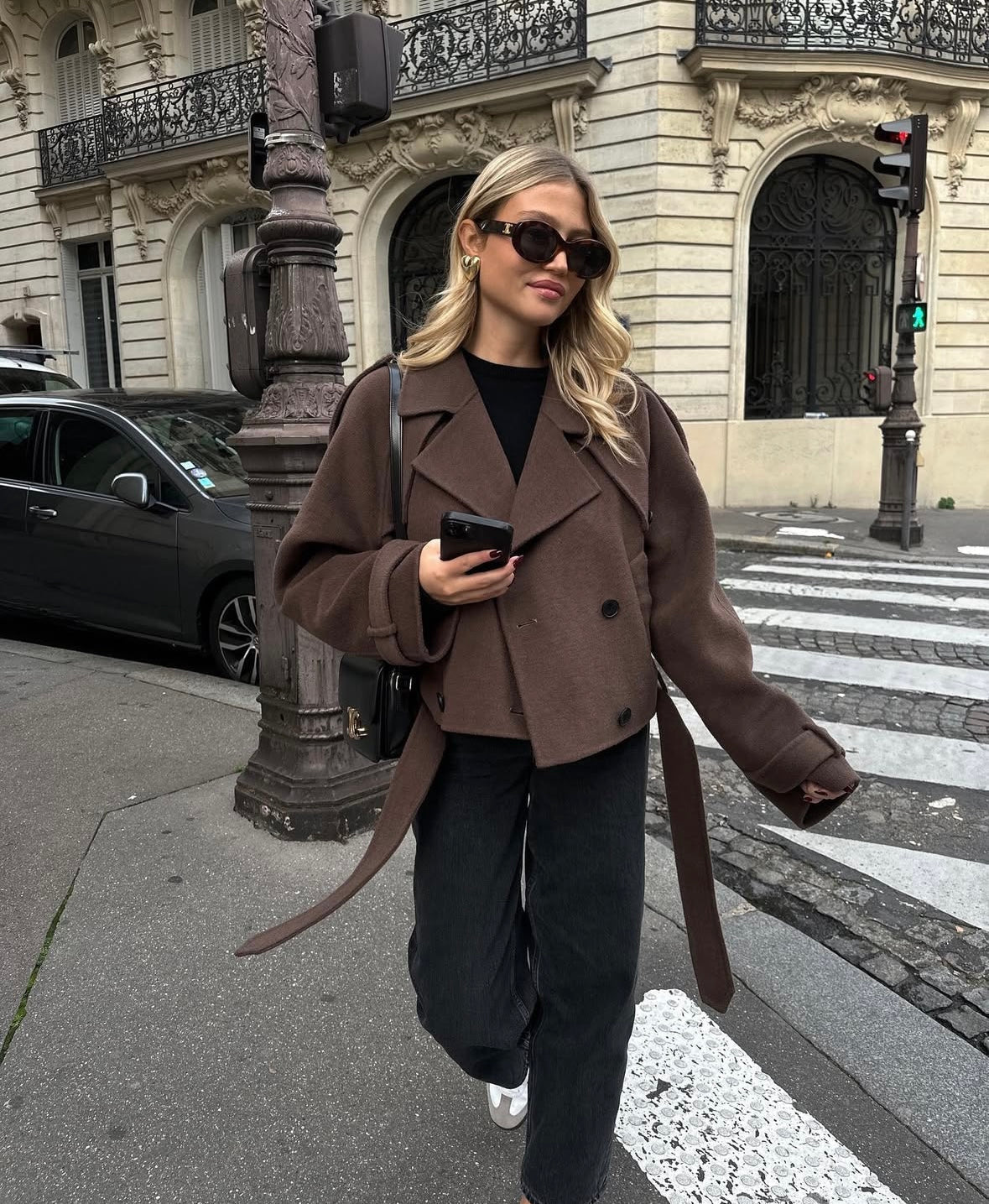 Ung kvinde i brun jakke og solbriller går på gaden i Paris. Hun holder en smartphone og bærer en sort taske. Elegant mode i bymiljø.