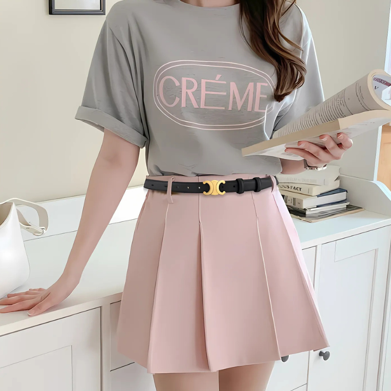 Kvinde iført grå "Crème" T-shirt og lyserød plisseret nederdel, holder en åben bog. Moderne mode, casual stil, trendy outfit.