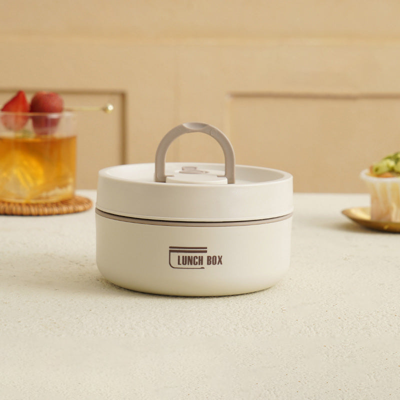 Hvid rund madkasse med håndtag på lys bordplade, ved siden af en drik med frugt og en cupcake. Perfekt til opbevaring af frokost og snacks.