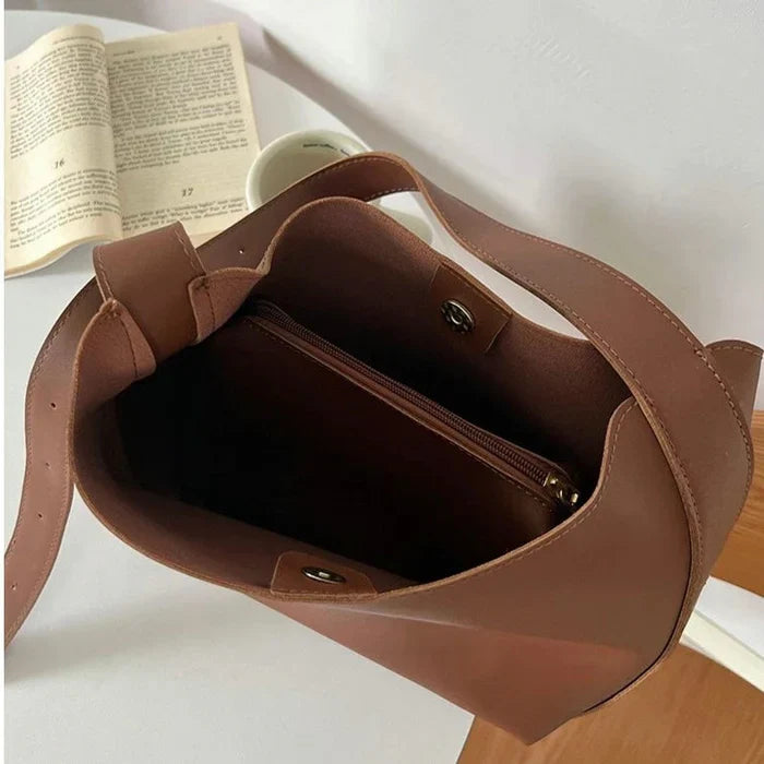 Brun lædertaske med åben top og indvendig lynlåslomme på hvidt bord, ved siden af en åben bog og en kaffekop. Stilfuldt og praktisk design.