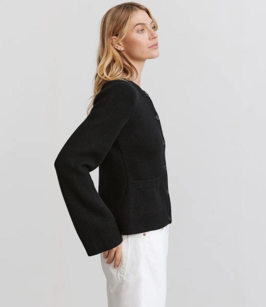 Kvinde iført sort cardigan og hvide bukser, set fra siden. Stilfuld mode, minimalistisk design, komfortabelt tøj, moderne look.