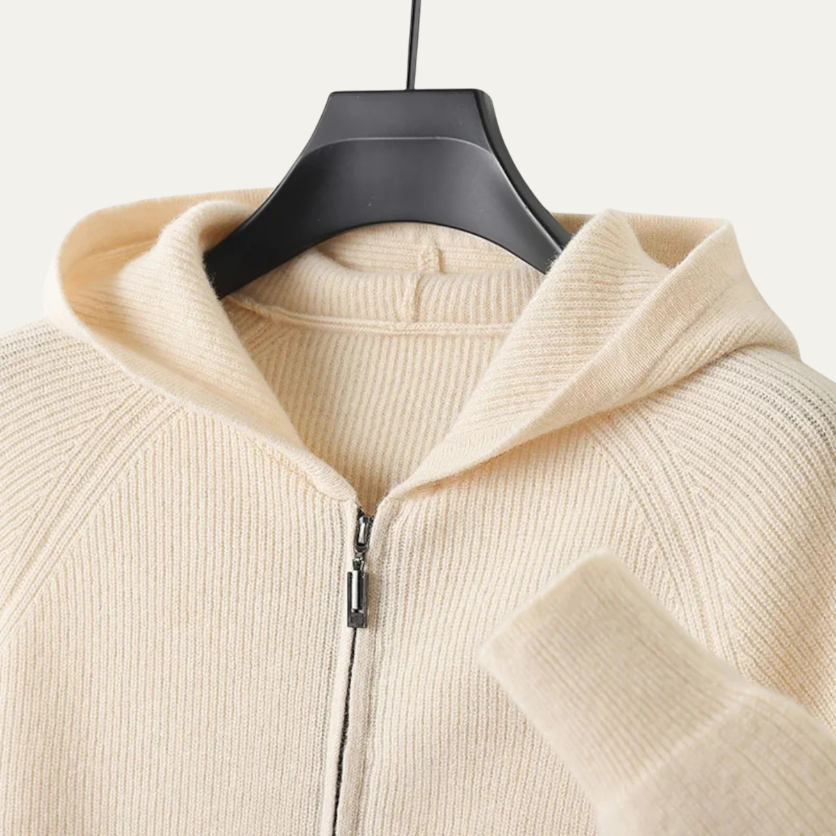 Beige hættetrøje i strik med lynlås, hængende på en sort bøjle. Komfortabelt og stilfuldt design, perfekt til afslappet mode og hverdagsbrug.