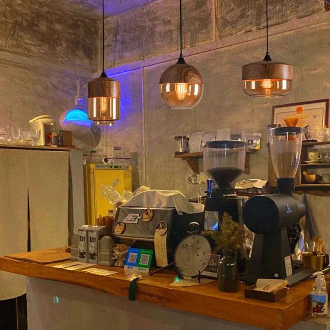 Hyggelig café med trælamper, espressomaskine og kaffekværn på trædisk. Industriel indretning, kaffebønner og dekorative elementer skaber stemning.