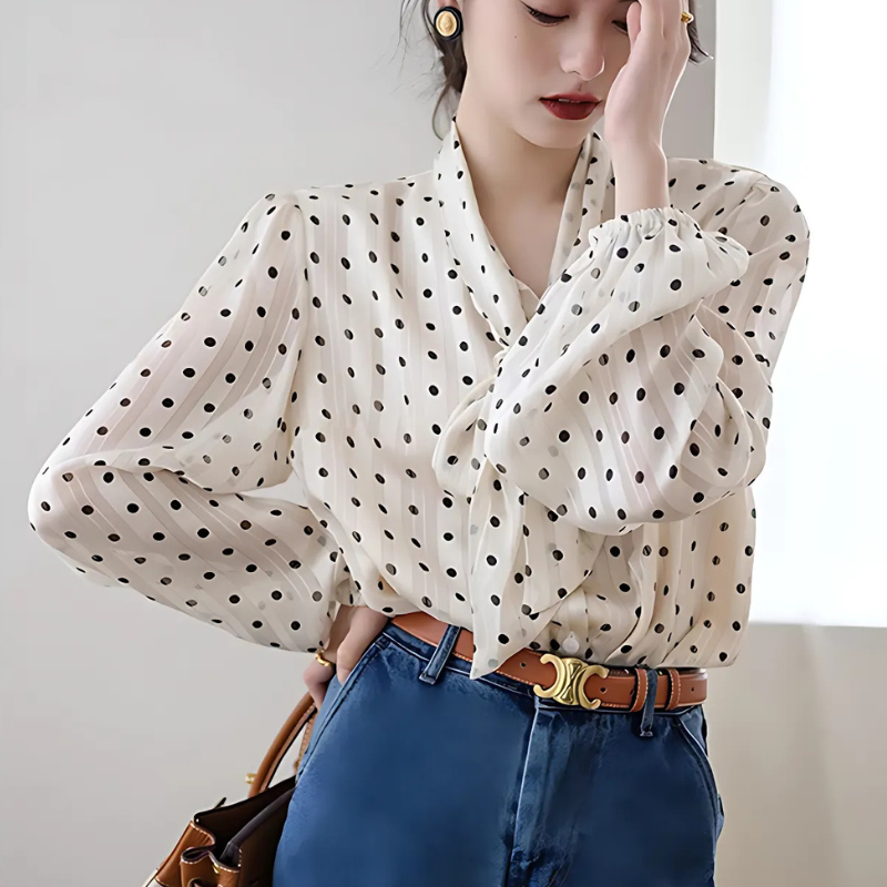 Kvinde iført hvid bluse med sorte prikker og blå jeans, holder en brun taske. Elegant og moderne mode, perfekt til hverdagsbrug.