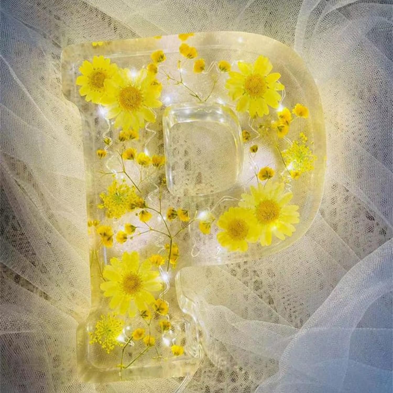 Bogstavet P i gennemsigtig harpiks med indlejrede gule blomster og LED-lys, placeret på hvidt tyl. Dekorativt, håndlavet design, unik boligindretning.