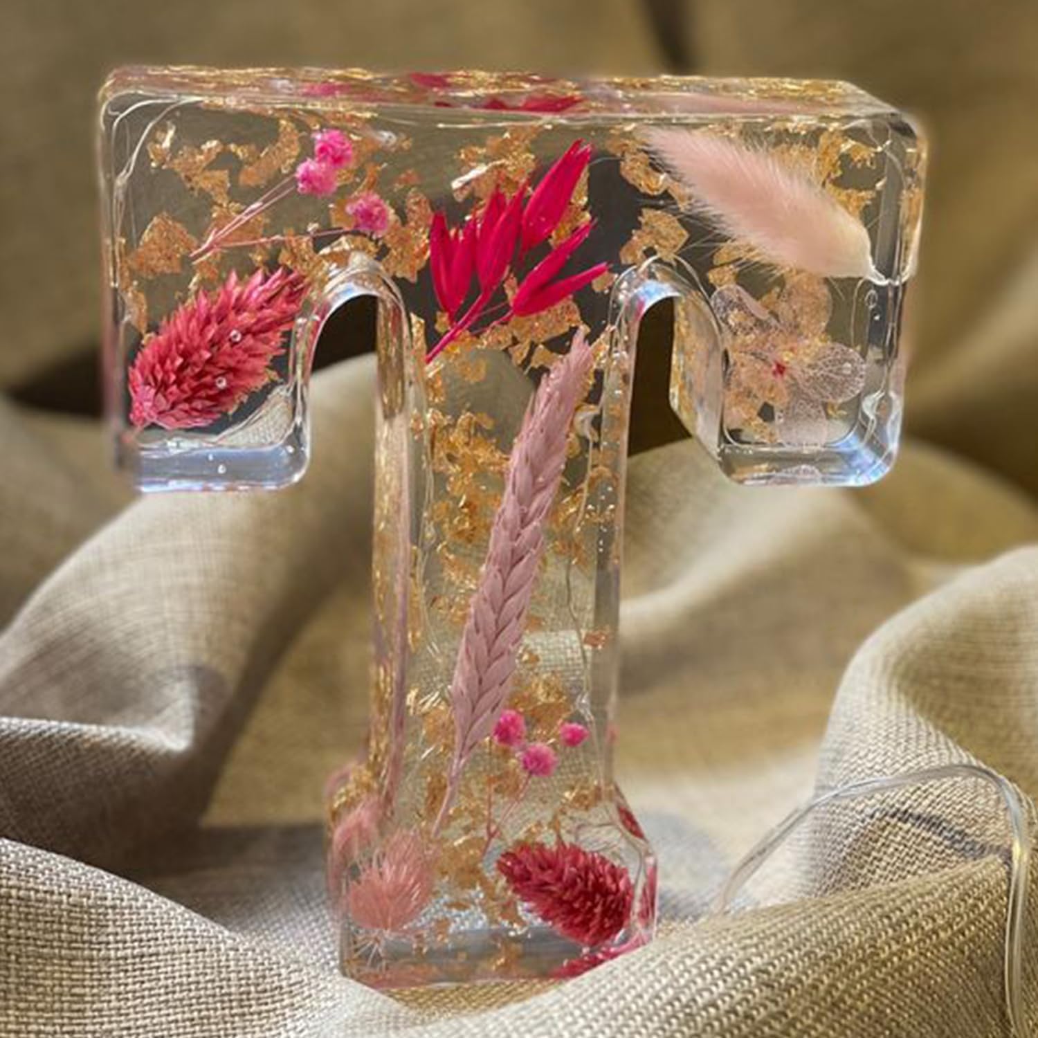 Bogstav T i klar harpiks med indlejrede tørrede blomster og blade, herunder lyserøde og røde nuancer, placeret på beige stofbaggrund.