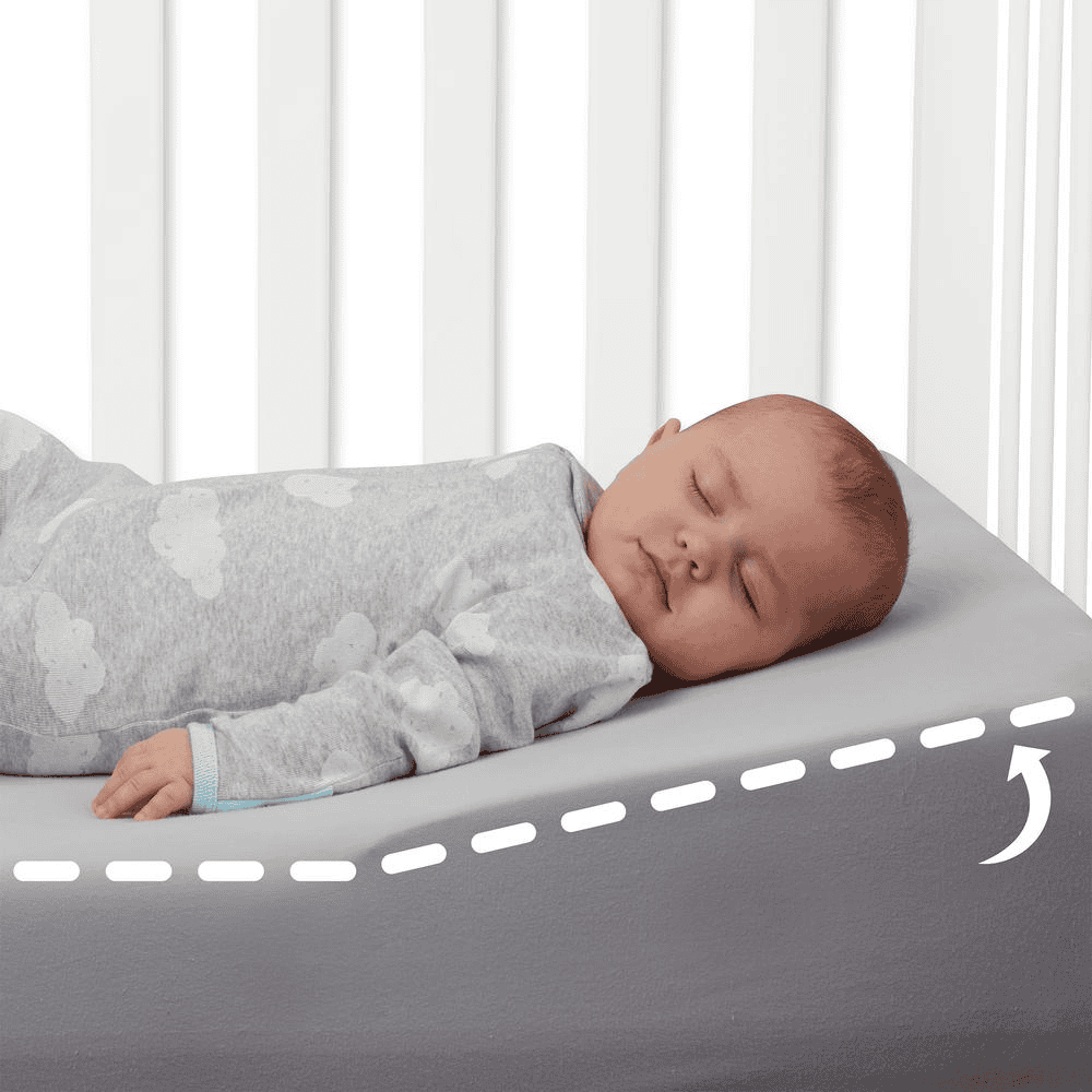 Baby sover på skrå madras i tremmeseng, iført grå sparkedragt med skyer. Ergonomisk designet for bedre søvn og komfort.