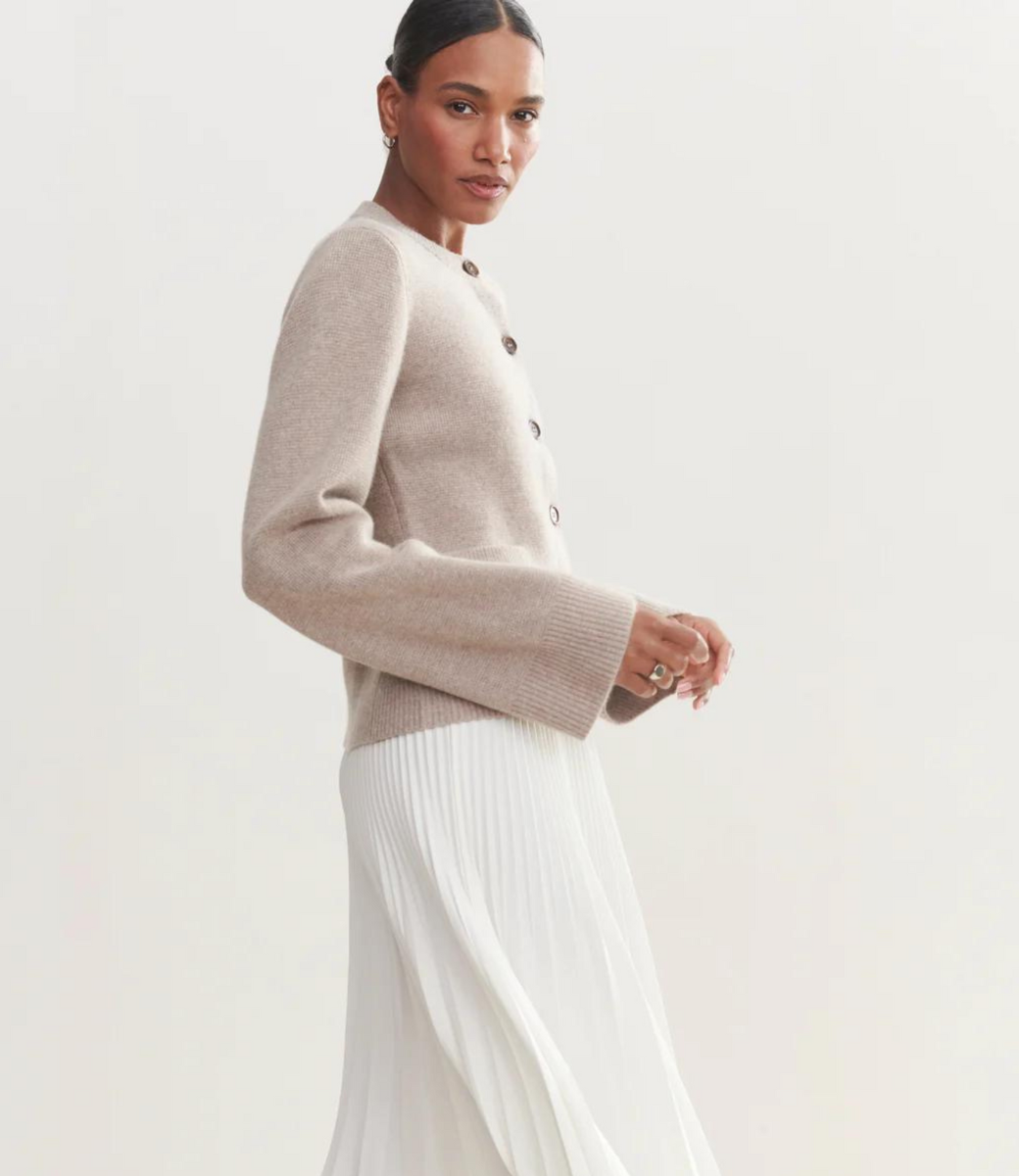 Kvinde iført beige cardigan og hvid plisseret nederdel, poserer mod lys baggrund. Elegant mode, minimalistisk stil, moderne kvindetøj.