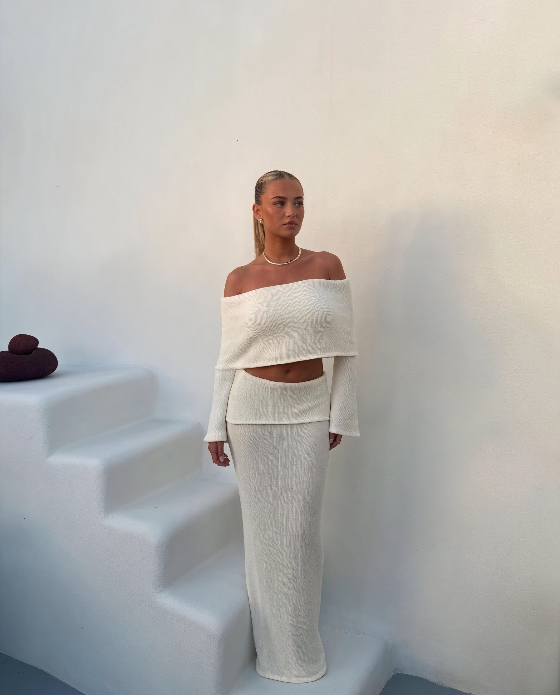 Kvinde i elegant, off-shoulder hvid kjole står ved hvide trapper. Minimalistisk mode, moderne design, stilfuldt look.