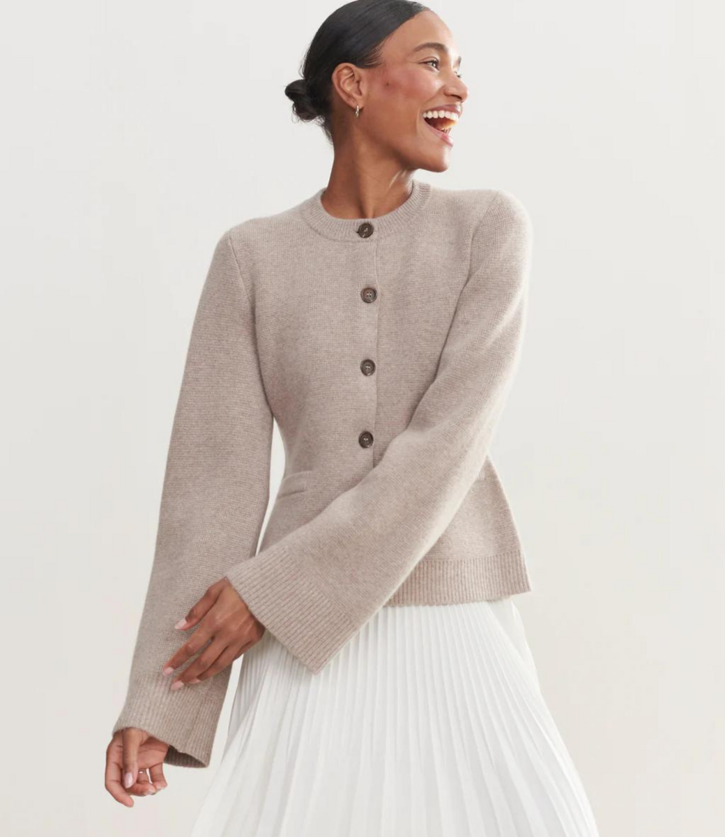 Kvinde iført beige cardigan med knapper og lange ærmer, kombineret med hvid plisseret nederdel. Elegant og moderne mode til kvinder.