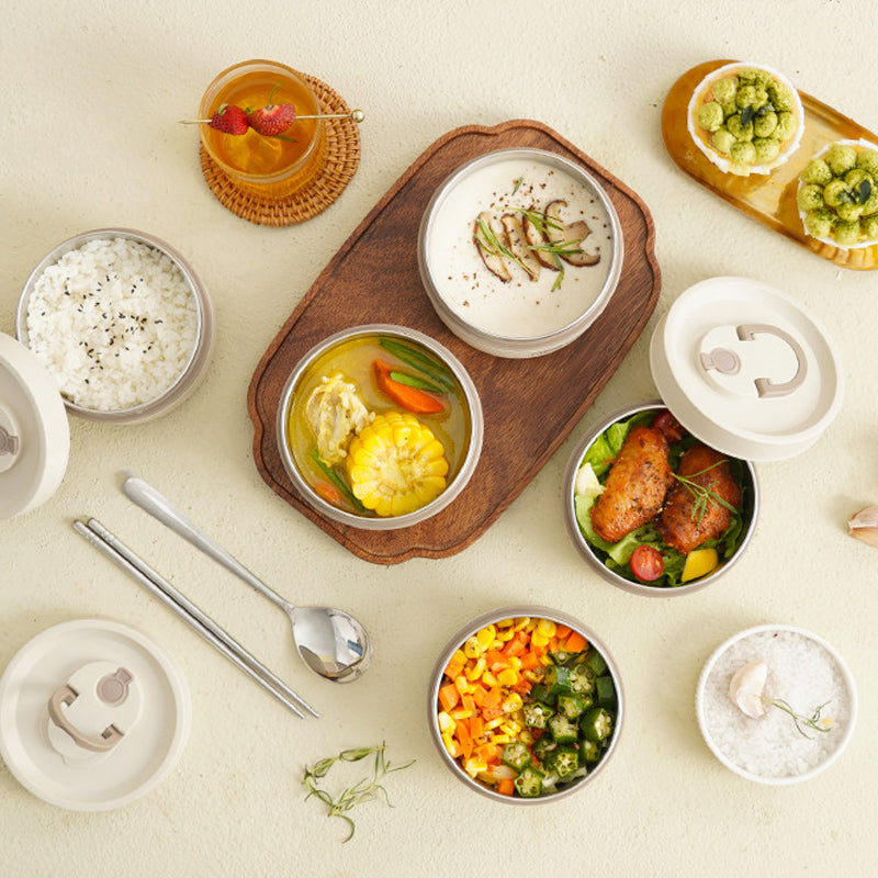 Frokostsæt med ris, suppe, grøntsager og kylling i runde beholdere på træbræt. Sund mad, bento-stil, serveret med te og bestik.