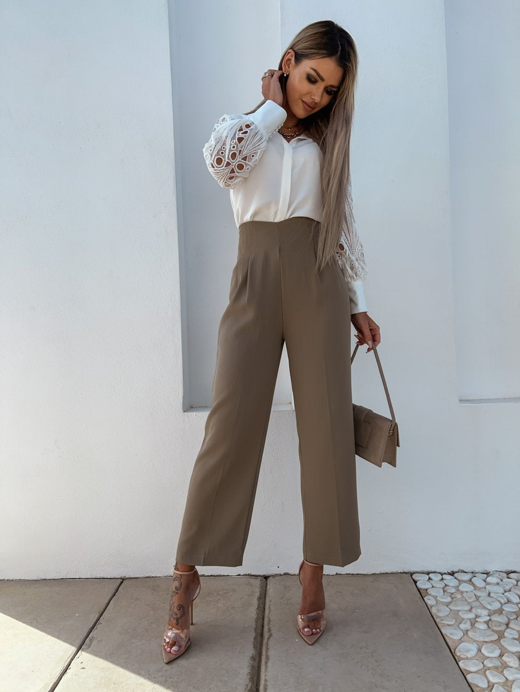 Kvinde i elegant outfit med hvid bluse og beige bukser, poserer mod hvid væg. Hun bærer en lille taske og har lange, lysebrune lokker.