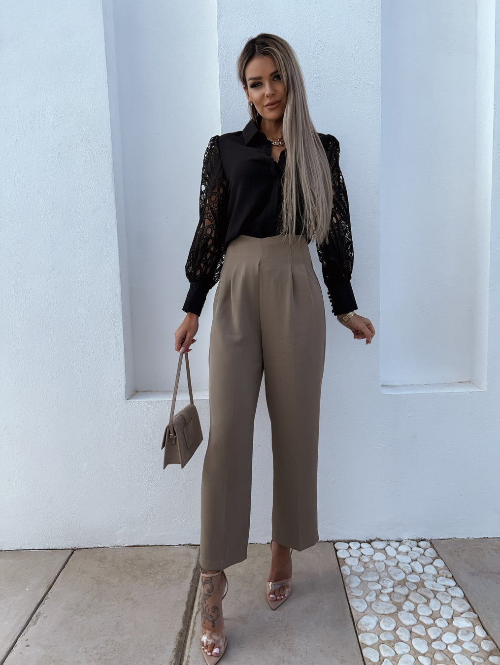 Kvinde i sort blondebluse og beige bukser, holder en lille taske. Står foran hvid væg. Mode, stilfuldt outfit, moderne kvindetøj.
