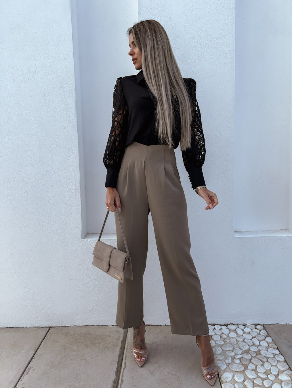 Kvinde i elegant outfit med sort blondebluse, beige bukser og matchende taske. Står mod hvid væg, iført stilfulde sandaler.