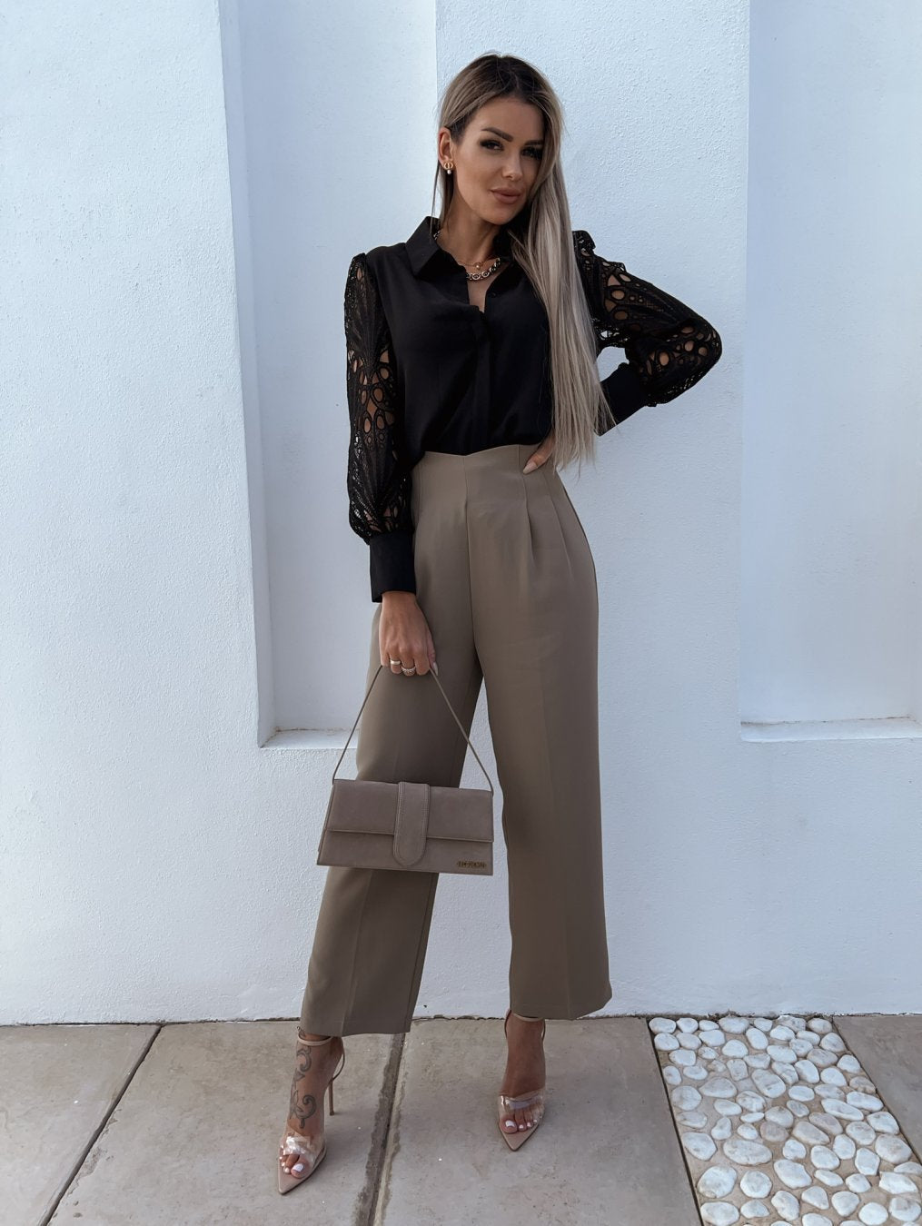 Kvinde i sort bluse med blondeærmer og beige bukser, holder en beige håndtaske. Står mod hvid væg, iført gennemsigtige hæle. Elegant mode.