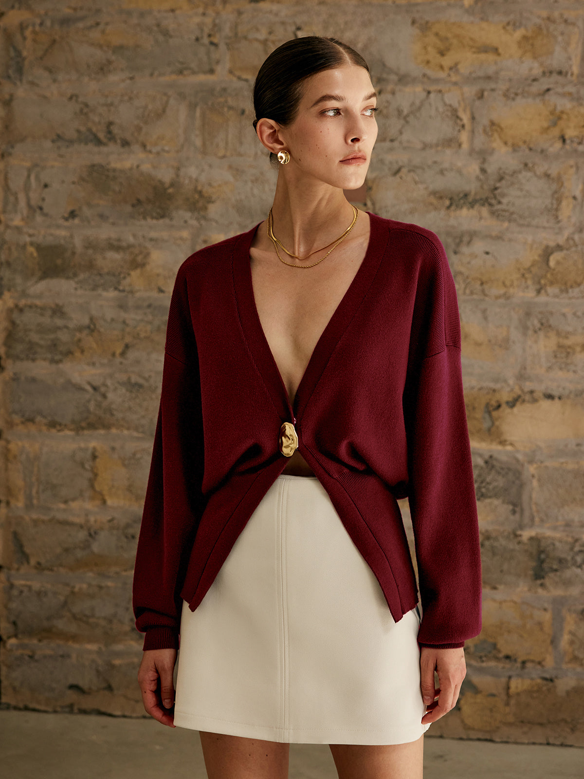 Kvinde iført bordeaux cardigan og hvid nederdel foran murstensvæg. Elegant mode, minimalistisk stil, moderne kvindetøj, efterårslook.