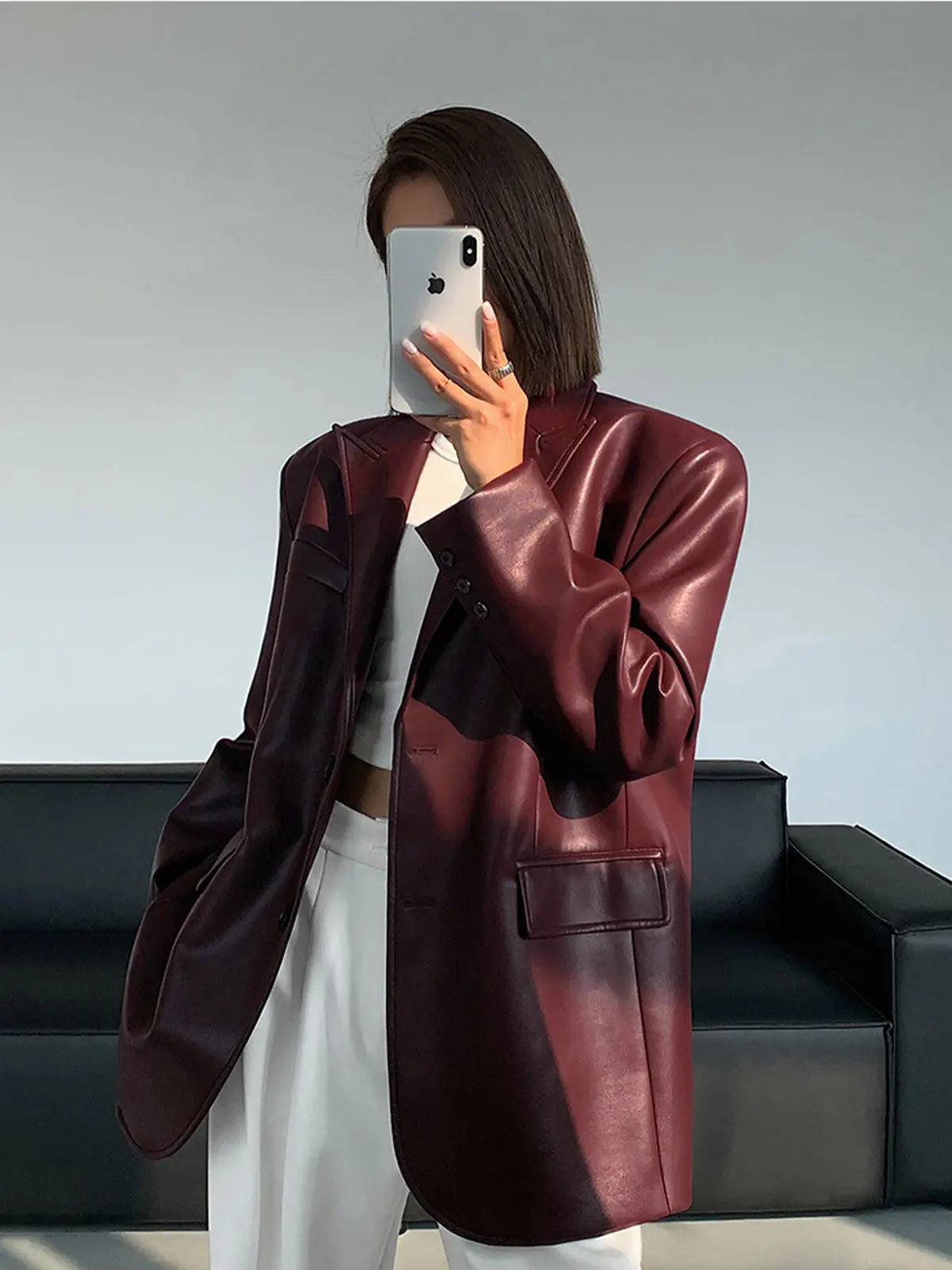Kvinde i bordeaux læderjakke tager selfie med smartphone, står foran sort sofa. Mode, stilfuldt outfit, moderne design, minimalistisk indretning.