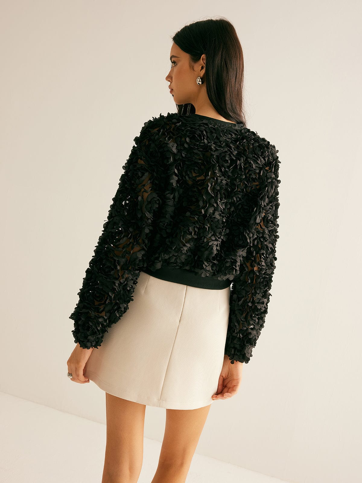 Kvinde iført sort blomstret jakke og beige nederdel, set bagfra. Elegant mode, moderne stil, tekstureret design, minimalistisk baggrund.