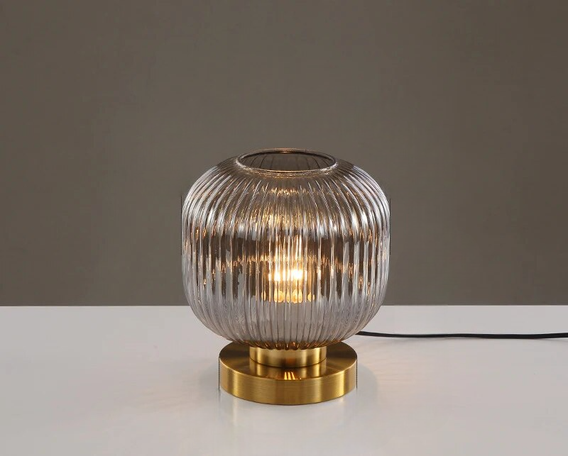 Rillet glasbordlampe med gylden base, moderne design, tændt pære, minimalistisk indretning, stilfuld belysning til stue eller kontor.