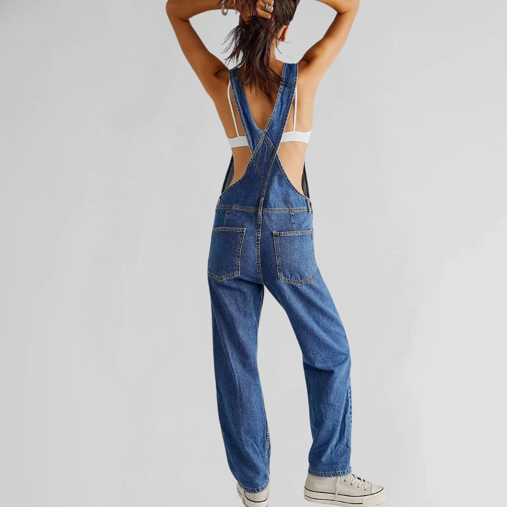 Kvinde iført blå denim overalls med krydsede stropper, hvid top og sneakers. Moderne afslappet mode, bagfra. Trendy streetwear look.