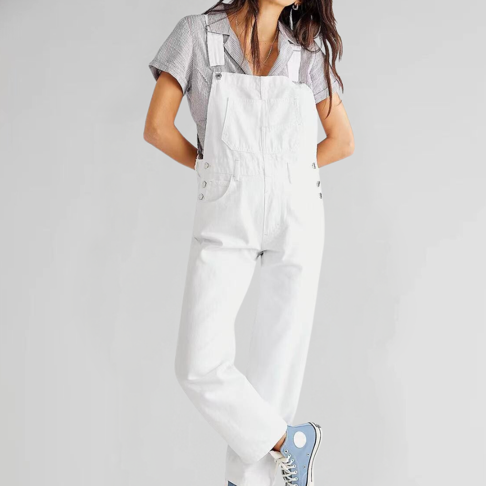 Ung kvinde iført hvid denim overalls og blå sneakers, poserer mod en grå baggrund. Moderne mode, casual stil, trendy outfit.