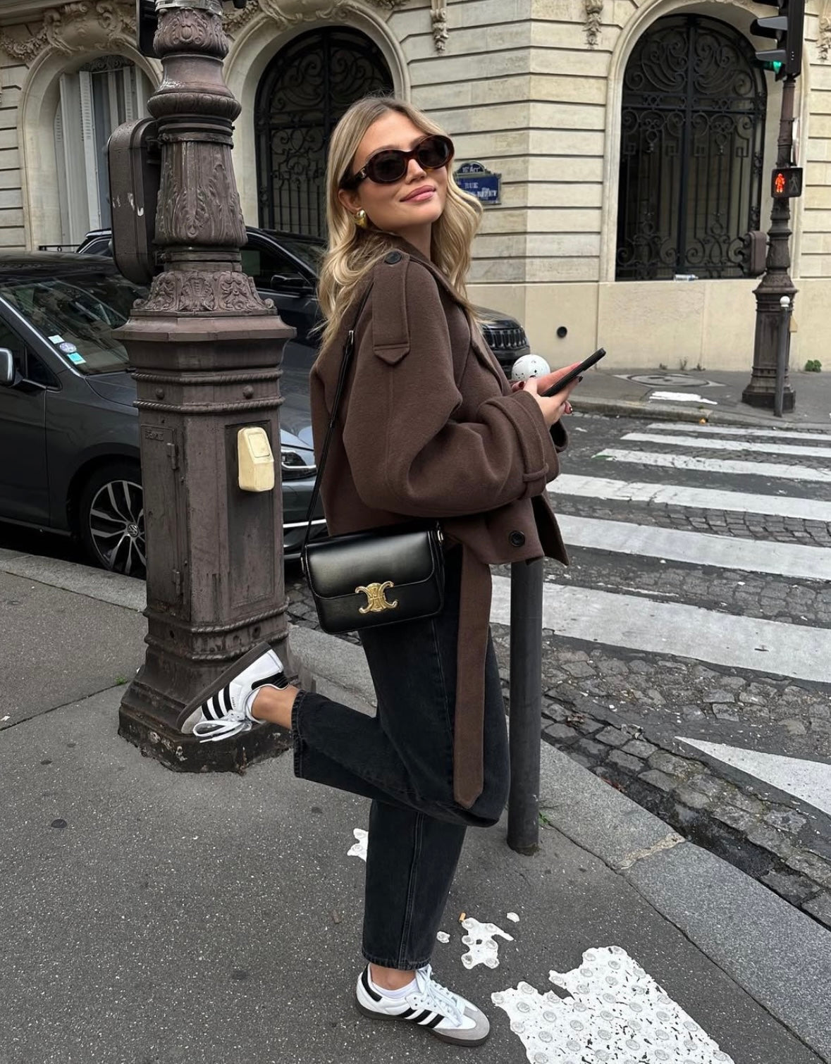 Ung kvinde i brunt frakke og sorte jeans står ved fodgængerfelt i Paris, bærer sort skuldertaske og solbriller, holder smartphone, smiler.