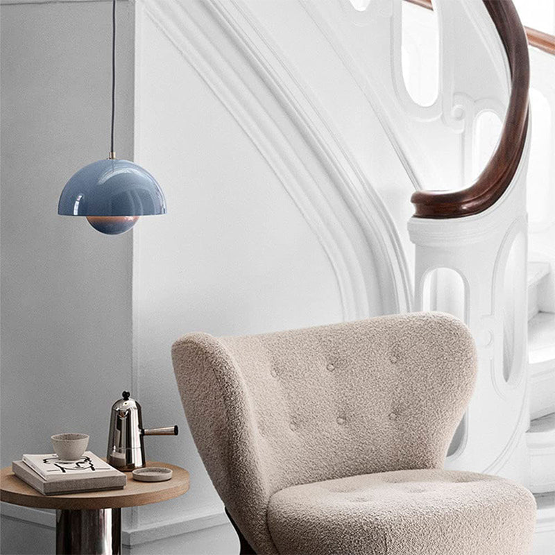 Blå pendellampe over beige loungestol ved hvid trappe. Moderne skandinavisk interiørdesign med kaffekande og bog på træbord.