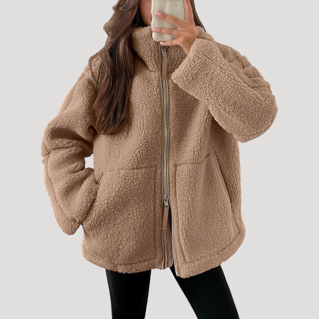 Kvinde iført beige teddyjakke med lynlås og lommer, poserer foran spejl. Vintermode, trendy overtøj, komfortabelt og stilfuldt design.