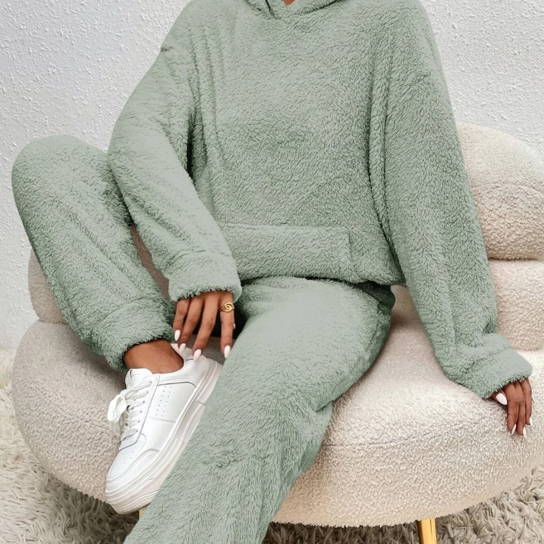 Kvinde iført lysegrønt teddyfleece sæt med hættetrøje og bukser, sidder i en beige stol. Komfortabelt loungewear, hvide sneakers, hyggelig stil.