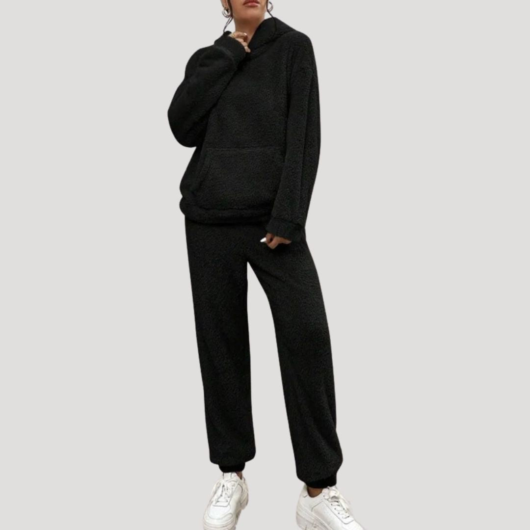 Kvinde iført sort fleece tracksuit med hætte og lommer, kombineret med hvide sneakers. Komfortabelt og stilfuldt outfit til afslappet brug.