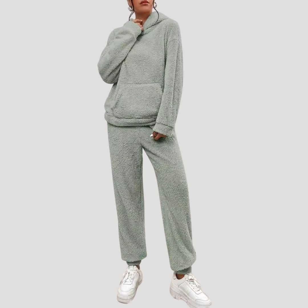 Kvinde iført grå fleece tracksuit med hætte og lommer, kombineret med hvide sneakers. Komfortabelt og stilfuldt loungewear til hverdagsbrug.