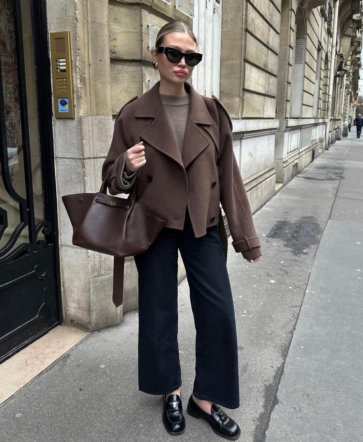 Ung kvinde i brunt jakkesæt, sorte bukser og solbriller, står på fortovet foran en klassisk bygning. Mode, street style, elegant look.