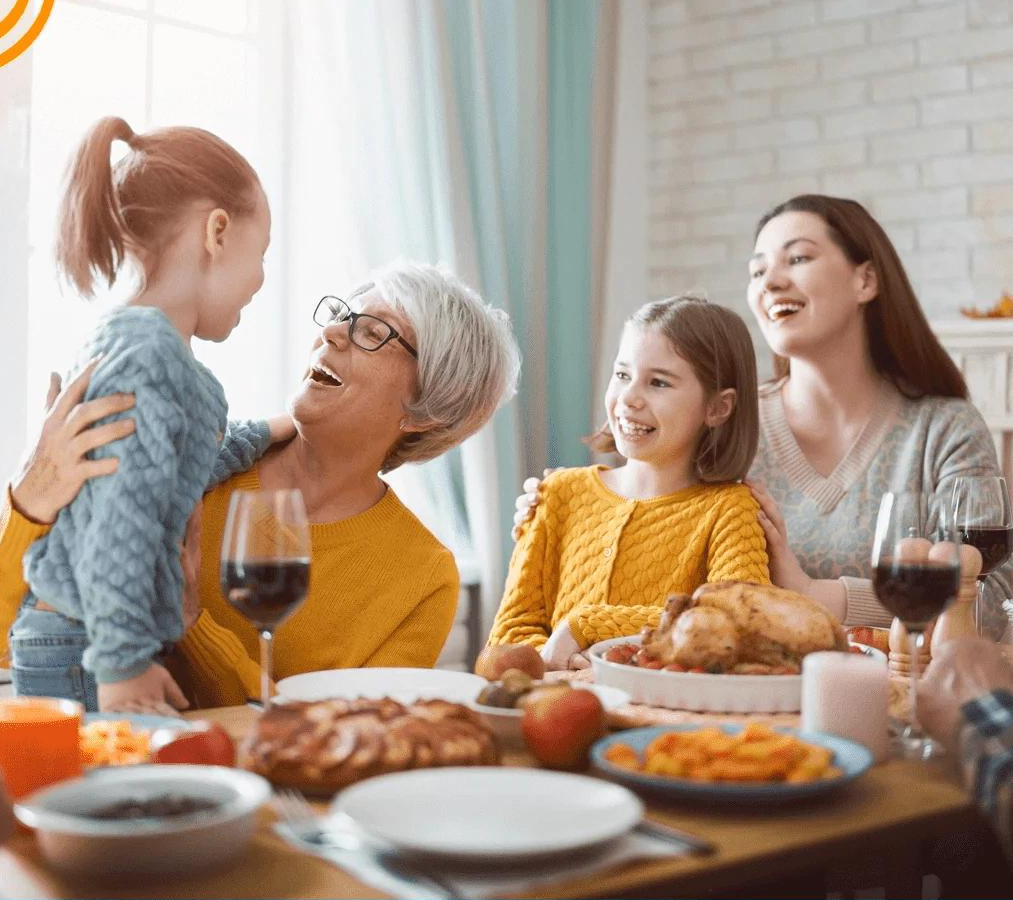 Familie hygger sig ved spisebordet med stegt kylling og vin. Tre generationer smiler og nyder måltidet i en lys stue. Familietid, middag, glæde.