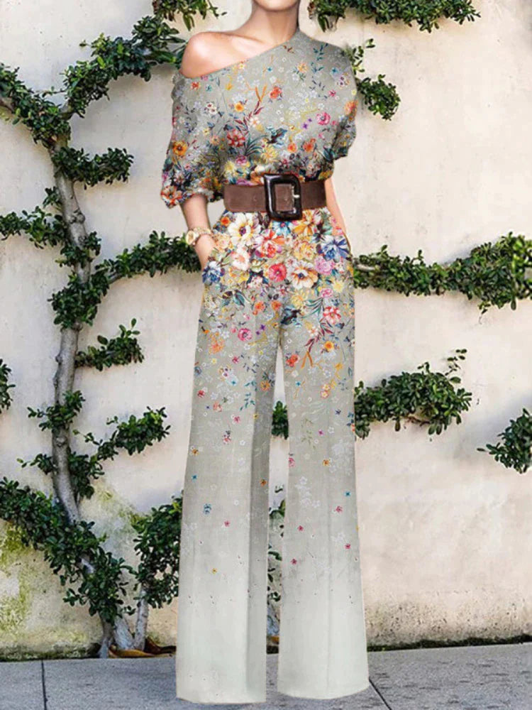 Kvinde i blomsterprintet buksedragt med bredt bælte, står foran væg med klatreplanter. Sommermode, elegant jumpsuit, trendy tøjstil.