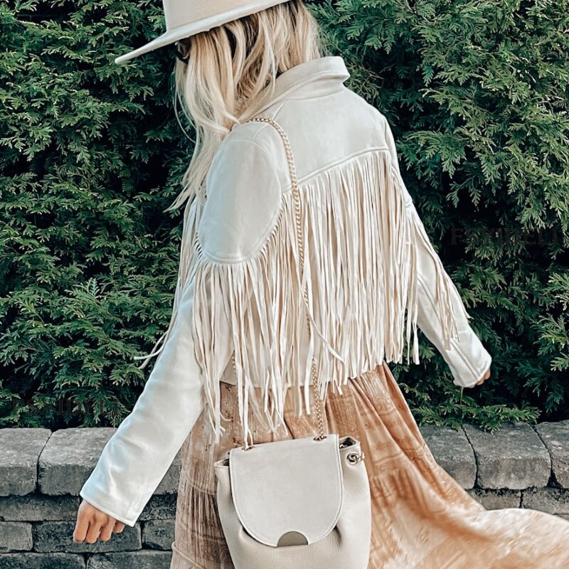 Kvindelig model i hvid frynsejakke og hat, bærer en beige taske. Står foran grønne buske. Mode, boho-stil, efterårsoutfit.