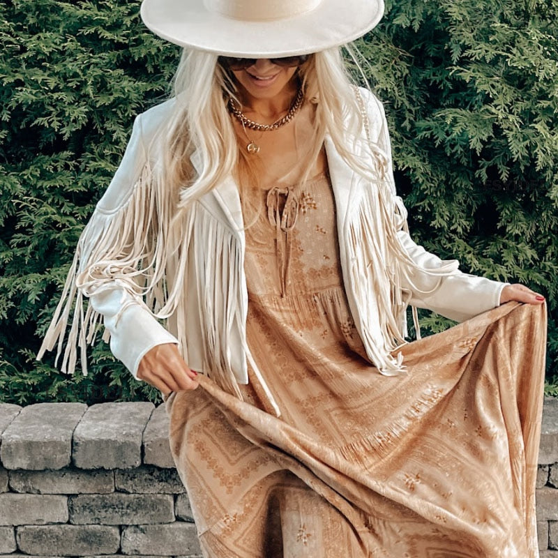 Kvindelig model i boho-kjole og hvid frynsejakke, iført bredskygget hat, poserer foran grønne buske. Boho-mode, sommerstil, kvindetøj.