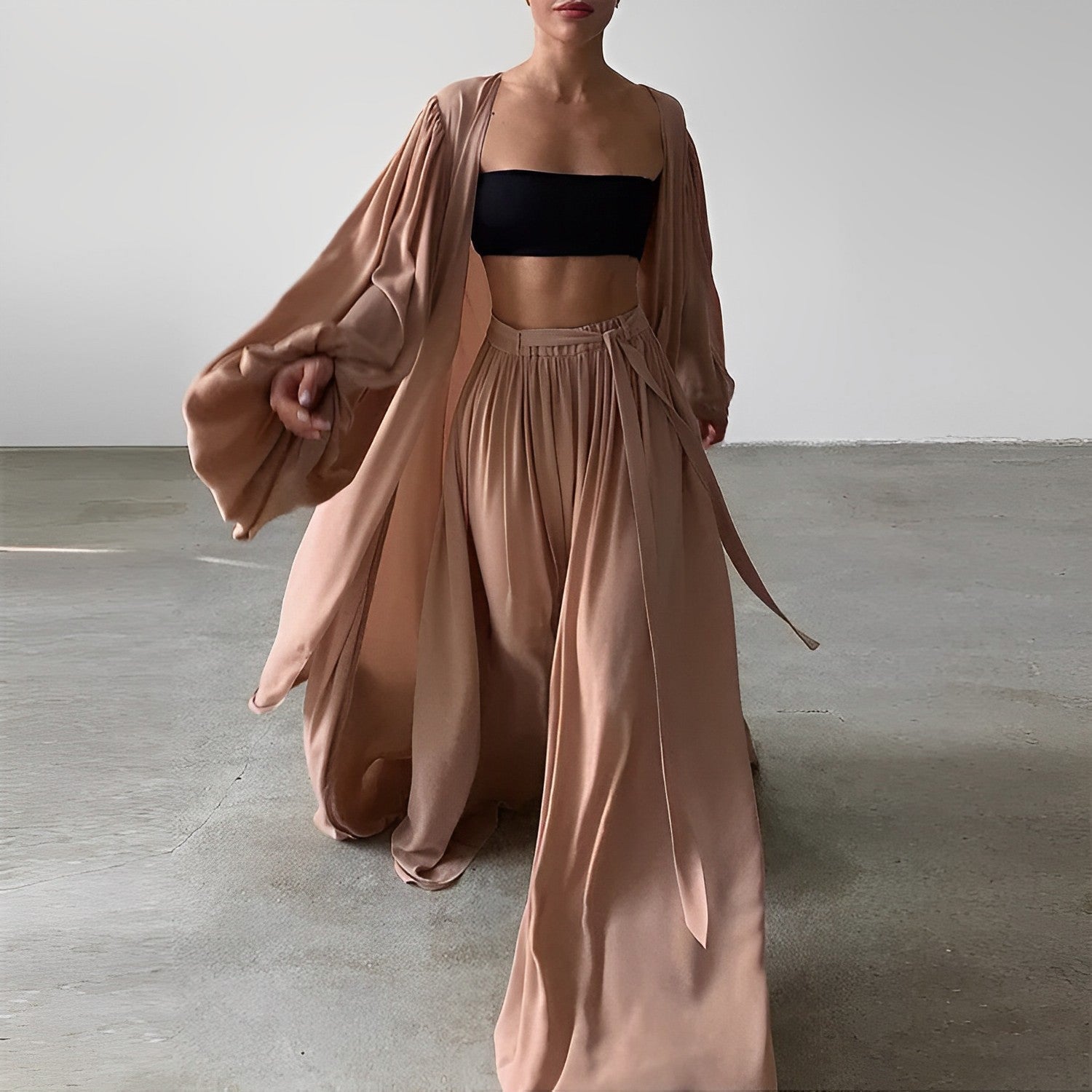 Kvinde iført elegant beige sæt med lang kimono og løse bukser, sort crop top. Moderne mode, minimalistisk stil, indendørs baggrund.
