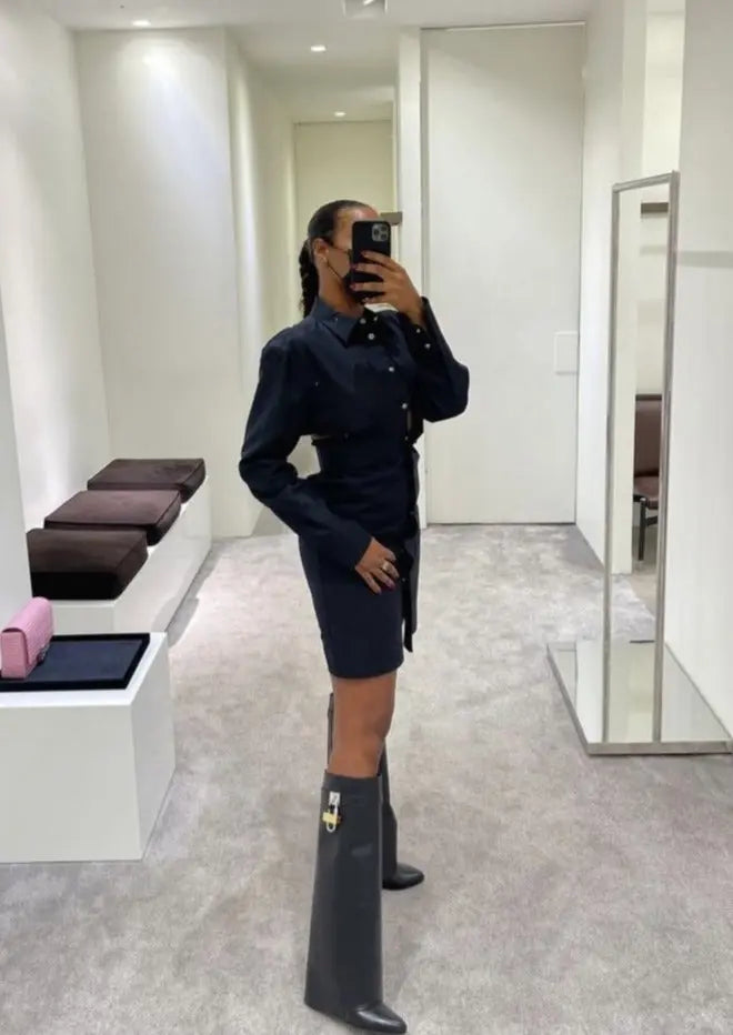 Kvinde i sort kjole og høje støvler tager selfie i moderne prøverum. Mode, shopping, stilfuldt outfit, spejl, minimalistisk indretning.