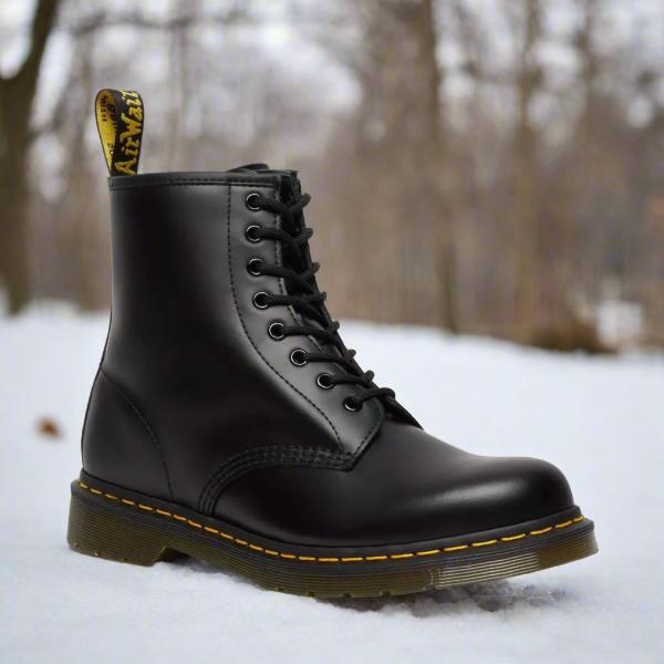 Sort Dr. Martens støvle i sneklædt skov, ikonisk design med gule syninger og snørebånd. Populær vinterfodtøj, robust og stilfuld.