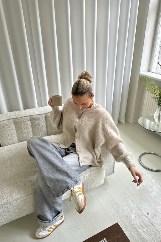 Kvinde i beige cardigan og grå jeans sidder på sofa med kaffekop. Moderne indretning, lyse gardiner, minimalistisk stil. Skandinavisk mode.