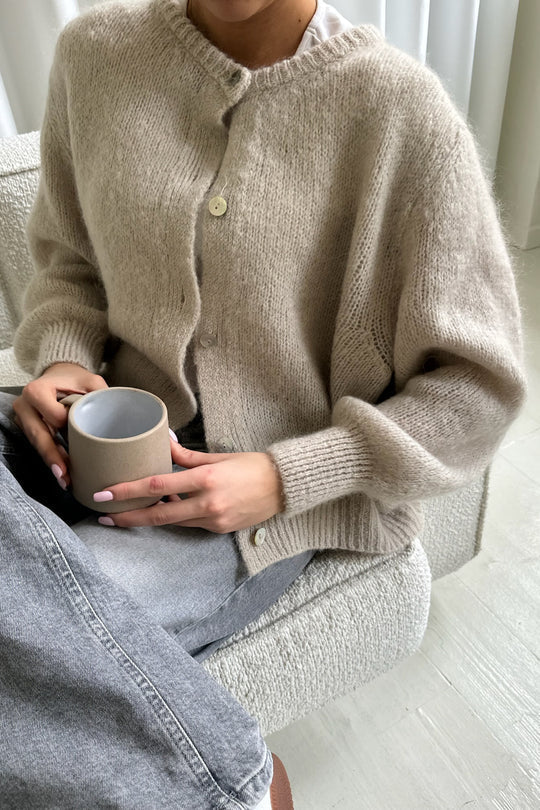 Kvinde iført beige striktrøje sidder på sofa med grå jeans, holder en keramisk kop. Hygge, mode, striktrøje, afslapning, skandinavisk stil.